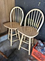 Two tall wooden stools with rounded backs and vertical slats, light wood, placed indoors on concrete floor near wall.