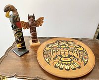 Photo showing three wood carved Indigenous totem figures and the round wall plate decorated with eagle design, all displayed on a wooden surface