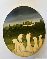 Front view of oval oil painting showing gaggle of seven white geese in grassy field with houses and trees in background.