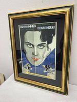 Angled photo of framed poster showing intense stylized female face with green eyes and red lips in Art Deco style.