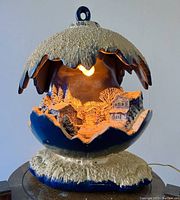 Front view showing illuminated interior snowy alpine village scene inside a ceramic Christmas ball with textured snow exterior.