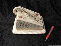Side view of the lidded cheese serving dish on its base plate, showing floral pattern and handle detail next to a pen for scale