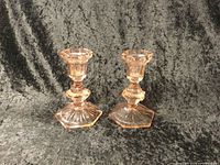 Photo shows two pink Depression glass candle holders with hexagonal bases and faceted stems against a dark fabric backdrop.