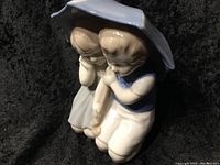 Porcelain figurine of boy and girl kneeling under umbrella, showing details and colors.
