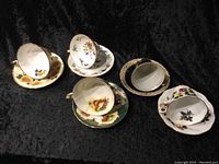 Five assorted fine bone china cups with matching floral saucers and gold trim, displayed tipped over and upright on black background.