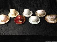 Seven fine bone china cups and saucers displayed flat on a black cloth background, showing different decorative designs and colors.