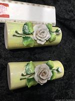 Photo showing both small yellow porcelain vases side by side with raised white rose and green leaves decoration on a black fabric background.