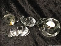 Photo showing all four items together on black fabric background: three solid glass animal figurines (rabbit, fish, pig) and one round glass candle holder with a tea light candle inside.