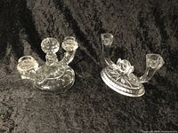 Photo of two clear depression glass candle holders on a black textured background showing intricate pressed glass patterns and shapes.