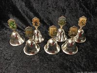 Eight silver plated bells arranged in two rows, each with a unique Hummel figurine plaque on top of the handle, showing colorful scenes.