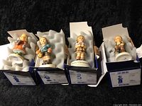 Four Hummel figurines displayed in original blue and white boxes, each with foam padding for protection. Figurines show children in traditional attire holding various items.