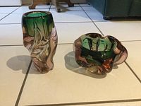 Photo showing two Murano glass pieces on a tiled floor: a heavy ashtray and a vase with green and pink colors.