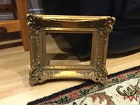Photo of an ornate gold gilded wood and plaster picture frame with floral and scroll decoration on slightly angled wooden floor with part of carpet visible