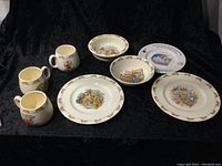 Photo showing seven pieces of Royal Doulton Bunnykins themed tableware including four small mugs, two shallow bowls, and one plate on a black background.