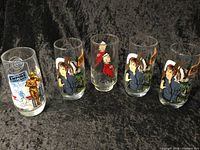 Five clear collectible glasses featuring Star Wars characters and Peanuts Star Wars theme arranged side by side on black velvet surface.