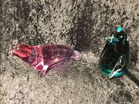 Pink seal and dark green penguin Venetian glass figurines on black textured surface.