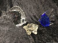 Three Venetian glass figurines displayed on a dark textured fabric background, including two fish (one clear 8-inch, one blue and clear 4-inch) and one clear frog figurine about 3.5-inch in size.