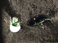 Photo shows two Venetian glass figurines on a dark textured background: one dolphin in dark green glass with clear fins, and one bird in white and green glass with clear wings.