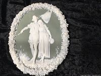 Oval Wedgewood style wall plaque with raised white figures of a man and woman in period dress, detailed floral border, and matte background. Mint condition.