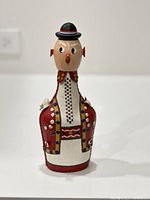 Front view of a 1970s vintage leather flask made in Yugoslavia, featuring a folk art figure wearing a detailed leather outfit with multicolored button decorations and a wooden head topped with a black hat.