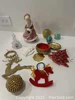 Overview of assorted Christmas ornaments and holiday figurines on display, showing various items including porcelain lady figurine, gold glitter reindeer, red felt rocking horse, and trinket box.