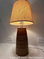 Front view of the ceramic table lamp illuminated, showing brown ribbed base and tan fabric shade