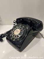 Front and side angle view of black rotary dial phone showing handset, coiled cord, and dial face with letters and numbers.