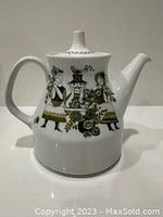 Front view of white ceramic tea pot showing stylized human figures and floral designs in green and black colors.