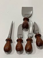 Full view of five cheese serving utensils with rosewood handles and stainless steel blades or forks, arranged side by side on a white background.