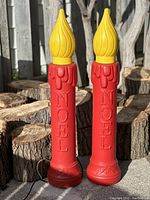 Pair of vintage blow mold candles with red body, yellow flame-shaped tops, embossed with 'NOEL'. Positioned outdoors on wood log background.