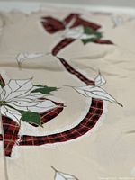 Close-up photo showing white poinsettia flowers with green leaves and red plaid ribbons printed on light beige cotton fabric, highlighting intricate gold outlines and lace details.