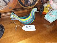 Side view of multicolor striped Murano glass bird figurine with tag numbered 97, placed on wooden surface.