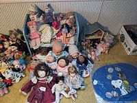 Wide view of the collection showing rag dolls, baby dolls, plush toys, small dollhouse, and blue doll bed filled with dolls and accessories