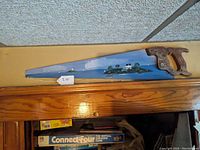 Full view of the antique hand painted saw blade with wooden handle lying flat showing the entire painted scene.