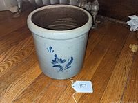Stoneware crock upright showing cobalt blue floral decoration on beige glazed surface, placed on hardwood floor with a lot tag labeled 107.