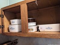 Wooden cabinet shelf displaying vintage CorningWare dishes in blue cornflower and spice of life patterns, including casserole dishes and a loaf pan.