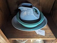Three Fiesta serving pieces stacked: blue pitcher on turquoise bowl on gray platter on wooden shelf.