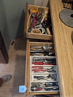 Two open wooden drawers showing the contents of kitchen utensils and flatware in white plastic drawer organizers. One drawer contains mostly stainless steel flatware with red and black handles, the other has a variety of kitchen tools such as peelers, wooden spoons, and mashers.