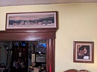 View of two vintage framed Norman Rockwell prints on canvas hung on a pale yellow wall above doorway. Large panoramic scene above door, smaller Santa Claus print to the right.