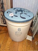 Front view of 6 gallon Uhl Pottery stoneware crock with blue painted wooden lid on hardwood floor next to radiator and framed artwork.