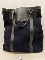 Front view of tote showing black woven fabric body, black leather top trim and dual handles