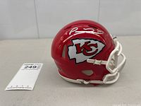 Full side view of red mini helmet with white facemask, Chiefs logo and silver autograph, lot tag visible
