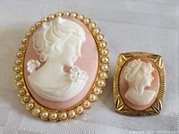 Photo showing two cameo brooches placed side by side on a white fabric background. Larger brooch is framed with faux pearls, smaller brooch has etched gold-tone frame.