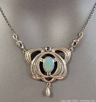 Close-up of pendant showing oval opal stone center with silver loops and small clear and black accent stones, plus a teardrop-shaped dangle.