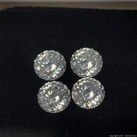Four round cut clear moissanite stones shown arranged on black surface, displaying brilliance and cuts.