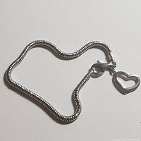 Silver snake chain bracelet laid out with a heart-shaped charm attached to the clasp, shown against a plain white background.