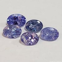 Five loose oval cut tanzanite gemstones shown on white background, varying shades of blue and violet, close comparison view