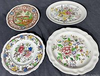 Four antique porcelain plates and platters shown on black fabric. Three have floral patterns, one has a scenic design with a house, trees, and border.