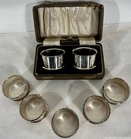 Pair of silver plate napkin rings inside a lined box, shown with five additional rings arranged around it on neutral background.