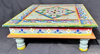 Side view of table showing floral and geometric hand painted patterns on wood, sitting on four short turned legs.